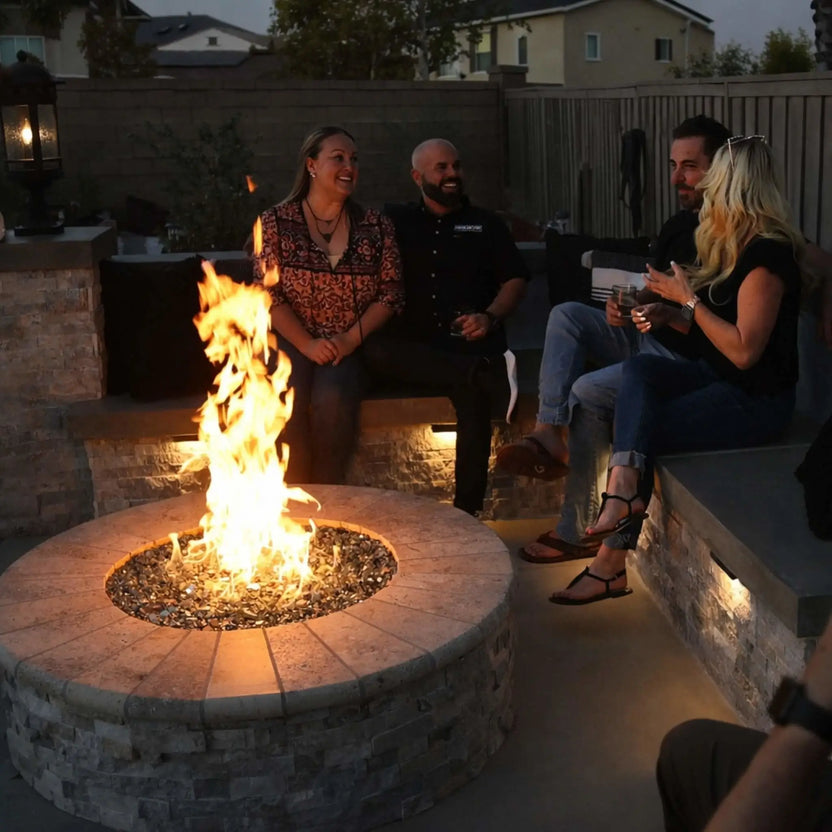 Evening backyard gathering around a round stone fire pit with a stainless steel burner pan and fire glass, where friends relax on built-in seating as warm flames illuminate the outdoor living space.