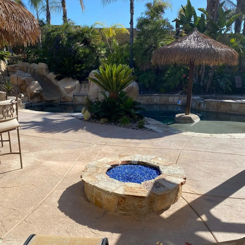 Low-profile round stone fire pit filled with cobalt blue fire glass, set on a stamped concrete patio beside a tropical-style backyard pool with lush palm trees, natural rock features, and a thatched umbrella creating a resort-inspired outdoor living space.