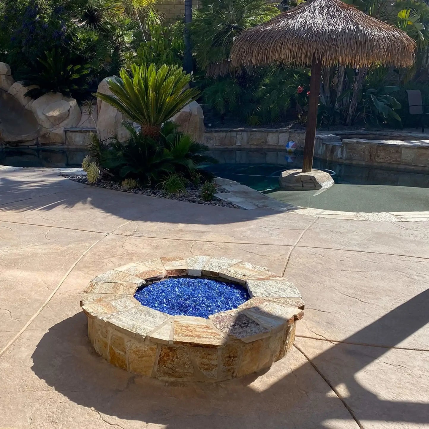 Circular outdoor fire pit constructed with stacked stone and capped with natural stone, featuring vivid blue fire glass at the center, positioned near a lagoon-style swimming pool surrounded by palm trees, tropical landscaping, and a shaded swim-up bar area.