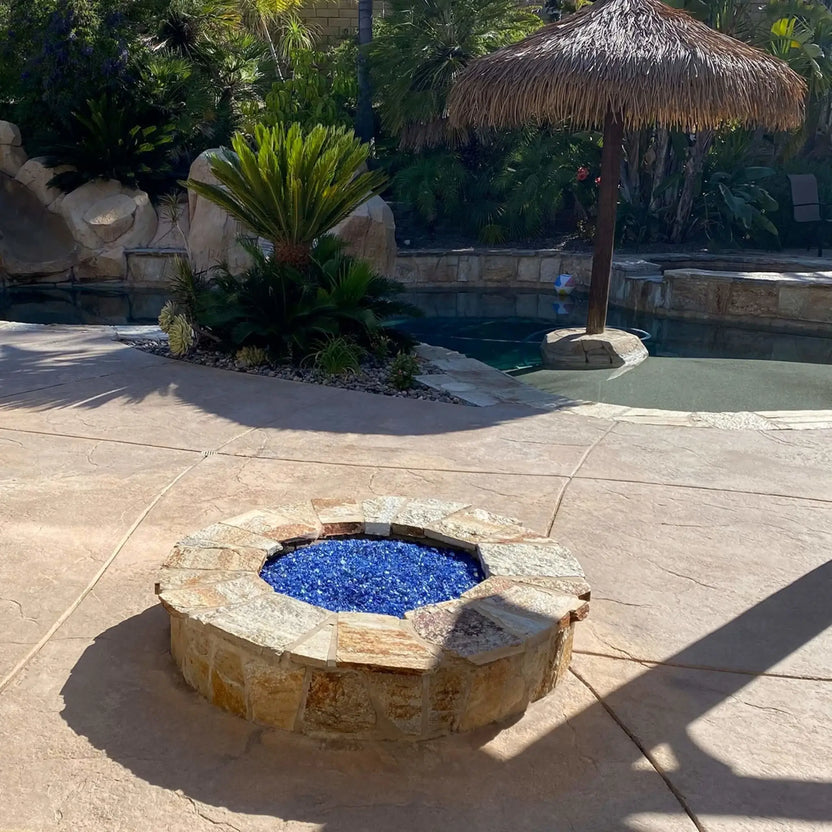 Circular outdoor fire pit constructed with stacked stone and capped with natural stone, featuring vivid blue fire glass at the center, positioned near a lagoon-style swimming pool surrounded by palm trees, tropical landscaping, and a shaded swim-up bar area.