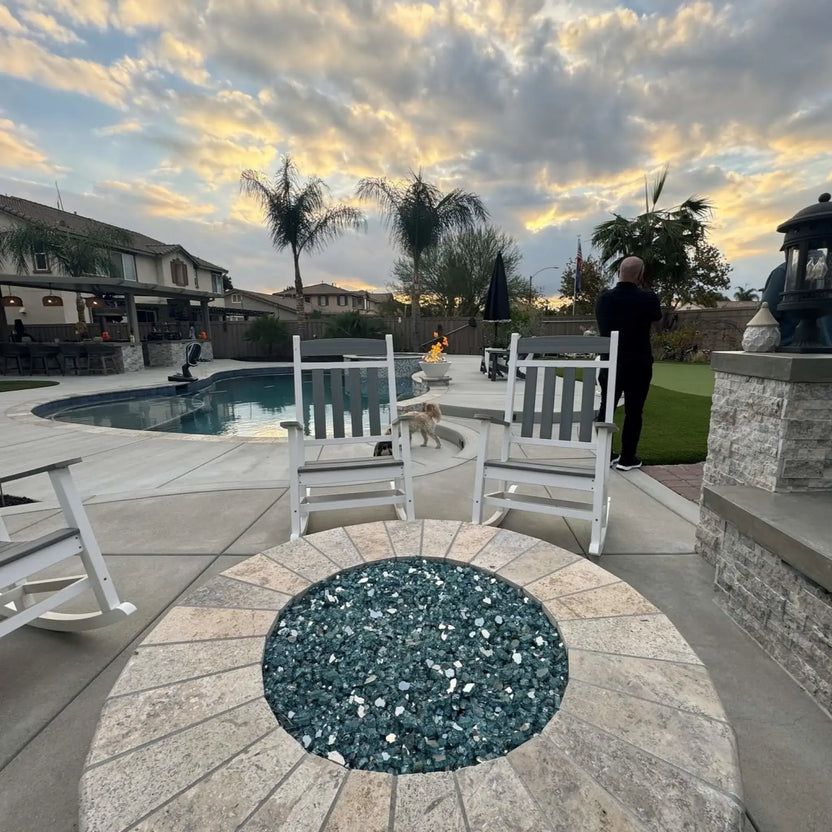 Modern round fire pit with stainless steel burner pan and reflective dark fire glass, centered on a concrete patio overlooking a backyard pool at sunset, with white rocking chairs, palm trees, and a landscaped lawn creating a relaxed evening gathering space.
