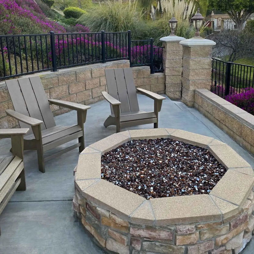 Octagonal stone fire pit filled with dark reflective fire glass, surrounded by Adirondack chairs on a concrete patio, backed by decorative stone walls, flowering shrubs, and soft landscape lighting for a cozy and inviting outdoor seating area.