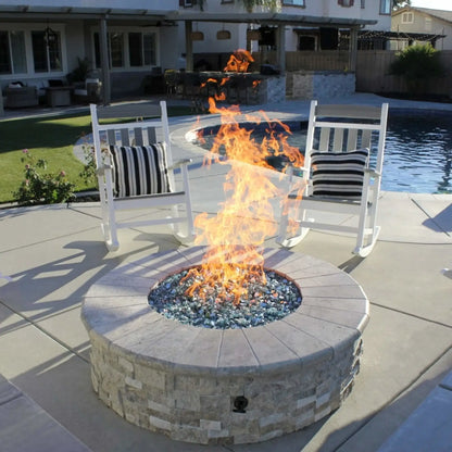 Round stone fire pit with a stainless steel flat burner pan producing tall, lively flames over blue fire glass, positioned on a modern poolside patio with white rocking chairs, a built-in outdoor kitchen, and a swimming pool in a landscaped backyard setting.