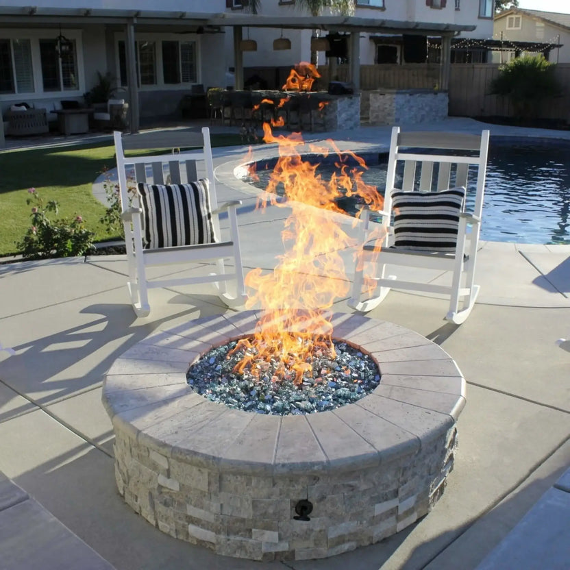 Round stone fire pit with a stainless steel flat burner pan producing tall, lively flames over blue fire glass, positioned on a modern poolside patio with white rocking chairs, a built-in outdoor kitchen, and a swimming pool in a landscaped backyard setting.
