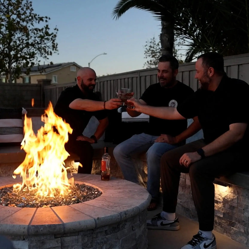Group of friends gathered around a circular stone fire pit at dusk, raising glasses beside a bright gas flame burning through decorative fire glass, creating a warm and inviting outdoor living space with built-in seating and accent lighting.