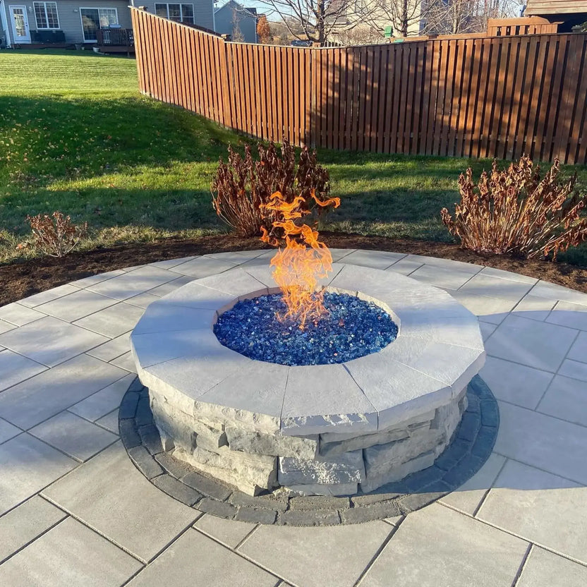 Custom round fire pit with a stainless steel burner pan and blue fire glass centerpiece, set on a paved patio surrounded by grass and a wooden privacy fence, showcasing steady flames and a clean, contemporary backyard fire feature design.