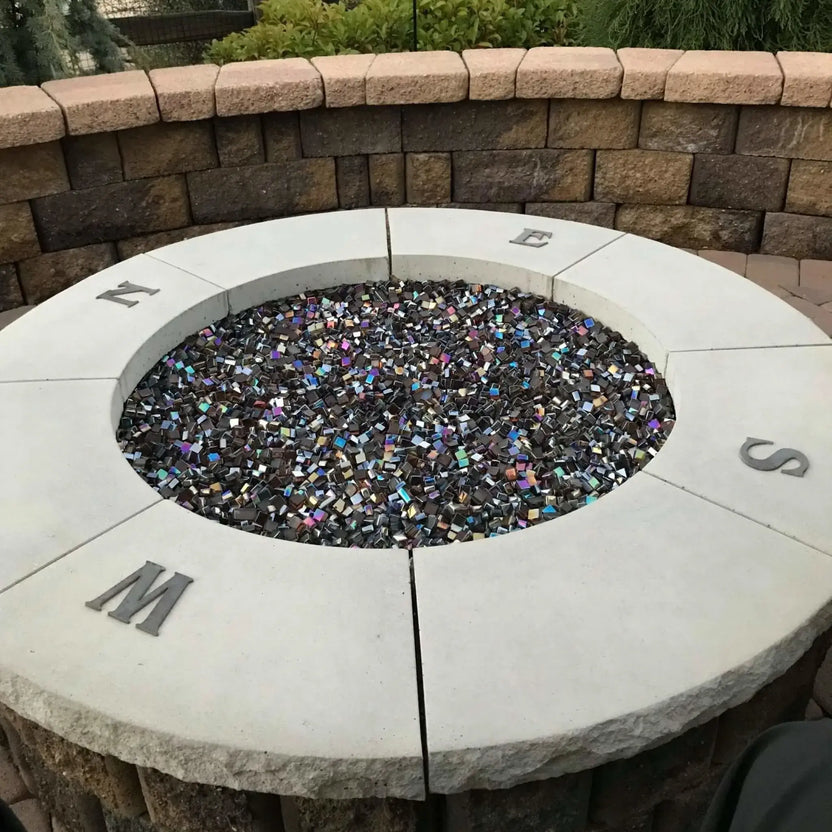 Round stone fire pit filled with dark reflective fire glass, shown unlit, set into a circular masonry surround with engraved compass direction markers, highlighting a clean and modern outdoor fire pit design.