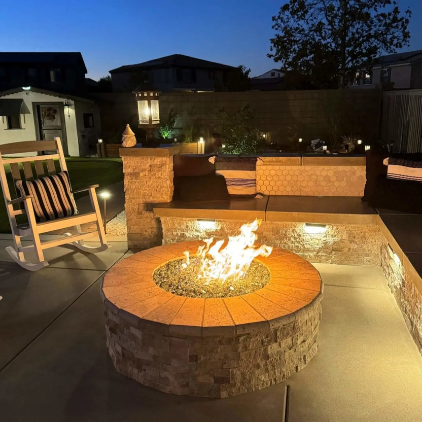 Nighttime backyard fire pit scene featuring a circular stone fire pit with glowing flames over fire glass, complemented by built-in seating, landscape lighting, and a white rocking chair, highlighting a cozy outdoor entertaining area after sunset.