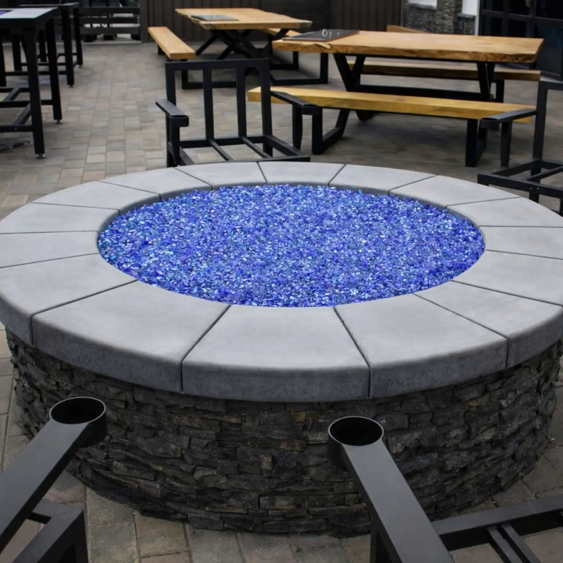 Round stone fire pit featuring a stainless steel flat burner pan filled with bright blue fire glass, set within a modern outdoor dining patio with wood picnic tables, metal chairs, and paved flooring for a clean commercial-style fire feature installation.