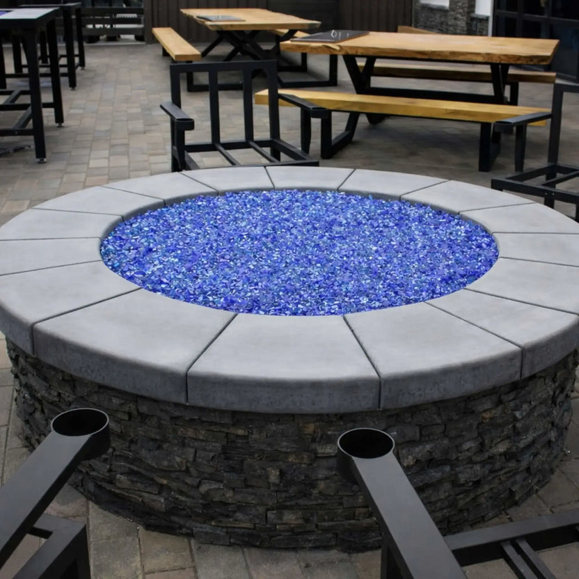 Round stone fire pit featuring a stainless steel flat burner pan filled with bright blue fire glass, set within a modern outdoor dining patio with wood picnic tables, metal chairs, and paved flooring for a clean commercial-style fire feature installation.