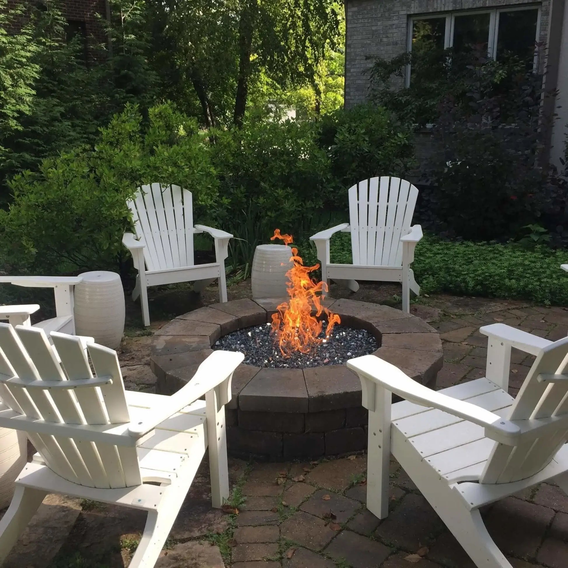 Outdoor circular fire pit burning bright orange flames over black fire glass, surrounded by white Adirondack chairs in a landscaped backyard patio, creating a warm and inviting gathering space.