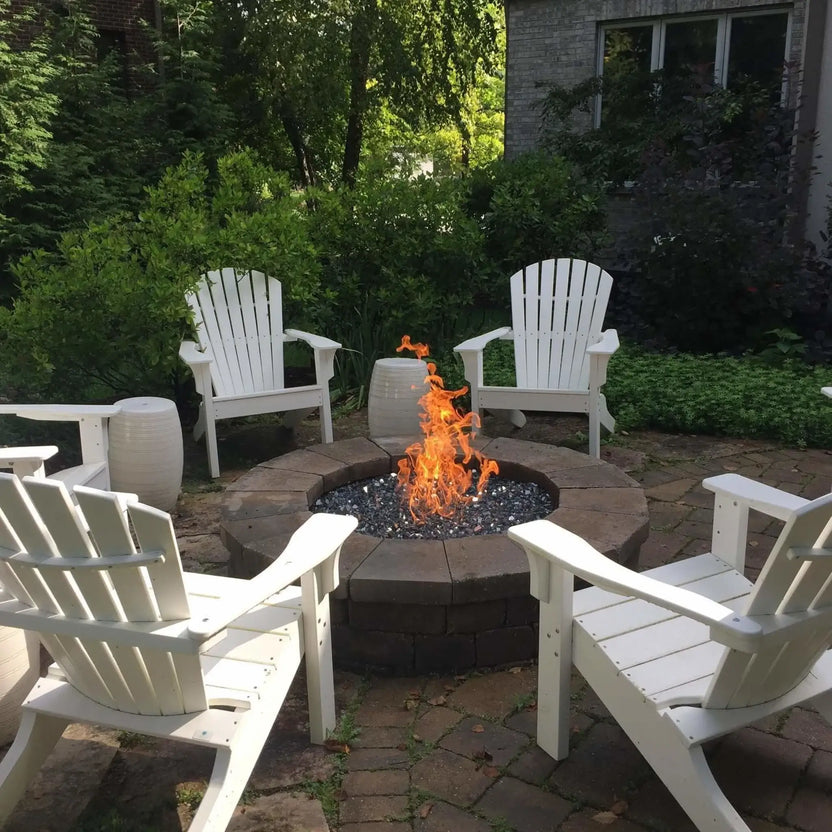 Outdoor circular fire pit burning bright orange flames over black fire glass, surrounded by white Adirondack chairs in a landscaped backyard patio, creating a warm and inviting gathering space.