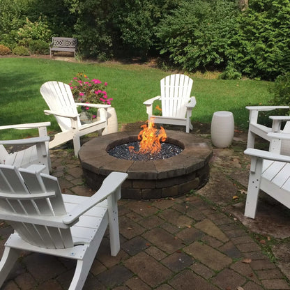 Backyard fire pit with a round stone surround and active flames rising from dark fire glass, positioned at the center of a seating area with white outdoor chairs and lush greenery.