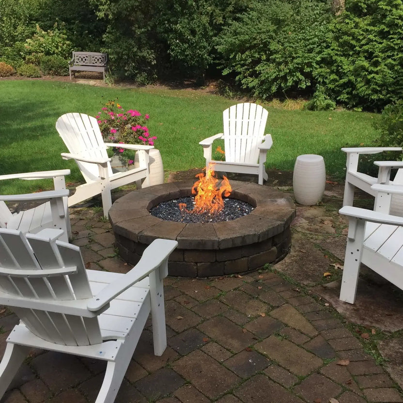 Backyard fire pit with a round stone surround and active flames rising from dark fire glass, positioned at the center of a seating area with white outdoor chairs and lush greenery.