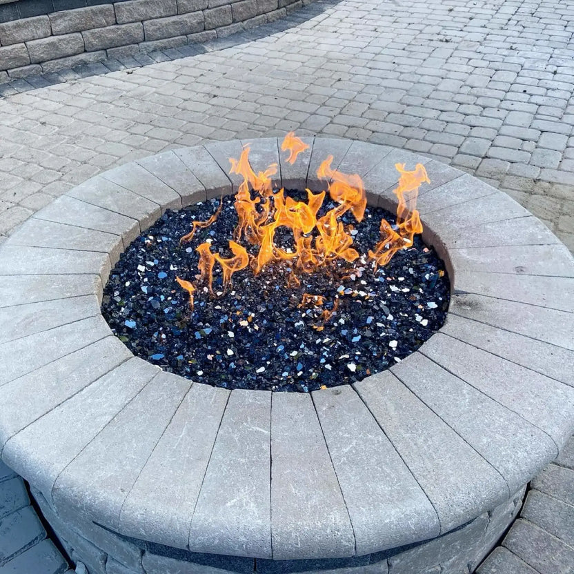 Close-up view of a circular stone fire pit featuring lively flames dancing across black fire glass, set into a paved patio surface that emphasizes the fire pit’s clean lines and modern outdoor styling.