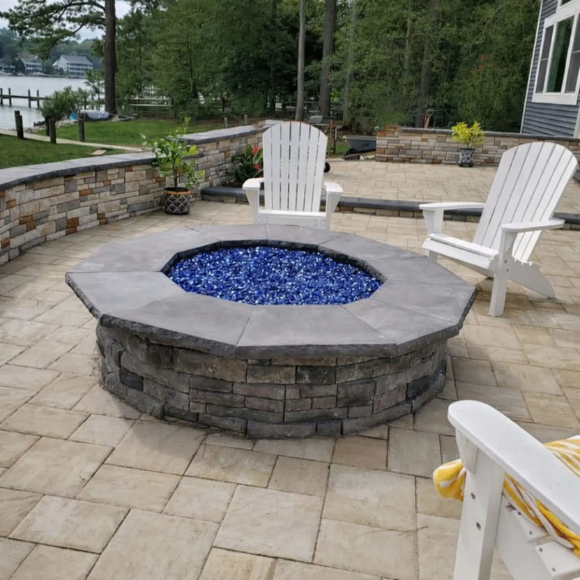 Round stone fire pit with blue fire glass fill installed on a paver patio, surrounded by white Adirondack chairs and low stone seating walls in a landscaped backyard setting.