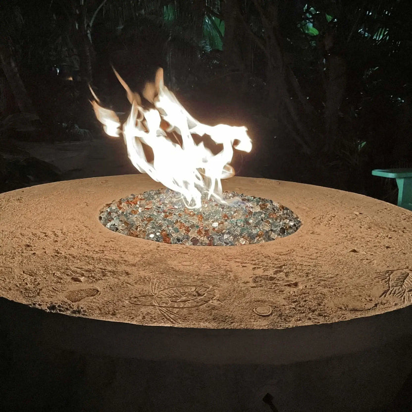 Nighttime view of a round outdoor fire pit burning bright flames over reflective fire glass, featuring a textured stone top and tropical landscaping in the background.