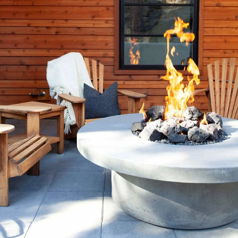 Modern round concrete fire pit with a stainless steel burner pan and natural lava rock, producing tall yellow flames in a cozy backyard seating area with wooden Adirondack chairs, side tables, and a warm wood-paneled exterior wall.