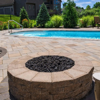 Circular stone fire pit with a stainless steel flat burner pan filled with black lava rock, positioned beside a swimming pool on a paved patio surrounded by landscaped greenery and residential backyard fencing.