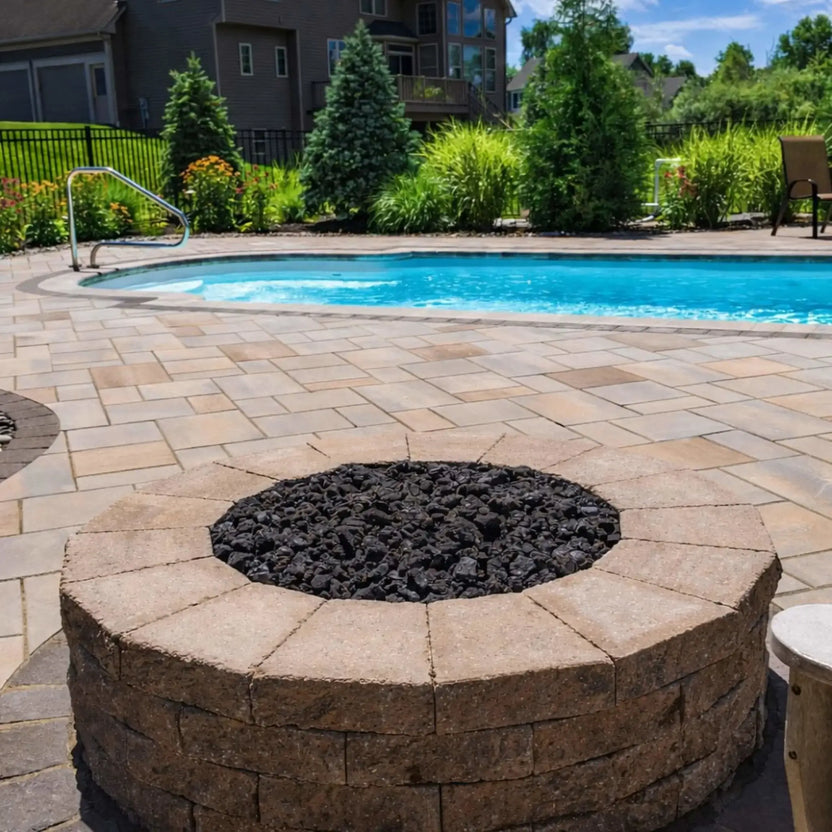 Circular stone fire pit with a stainless steel flat burner pan filled with black lava rock, positioned beside a swimming pool on a paved patio surrounded by landscaped greenery and residential backyard fencing.