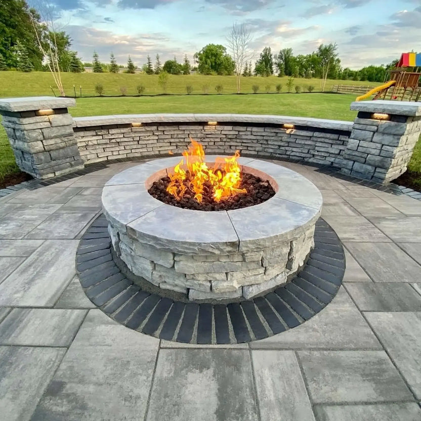 Large round masonry fire pit with a stainless steel burner pan and lava rock, actively burning with vibrant flames in an open outdoor gathering space featuring curved stone seating walls, wide paver flooring, and expansive green lawn views.