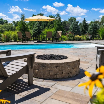 Round stone fire pit filled with black lava rock, positioned on a poolside paver patio with Adirondack chairs and a clear blue swimming pool in the background, surrounded by lush landscaping and outdoor lounge seating.