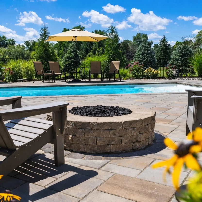 Round stone fire pit filled with black lava rock, positioned on a poolside paver patio with Adirondack chairs and a clear blue swimming pool in the background, surrounded by lush landscaping and outdoor lounge seating.