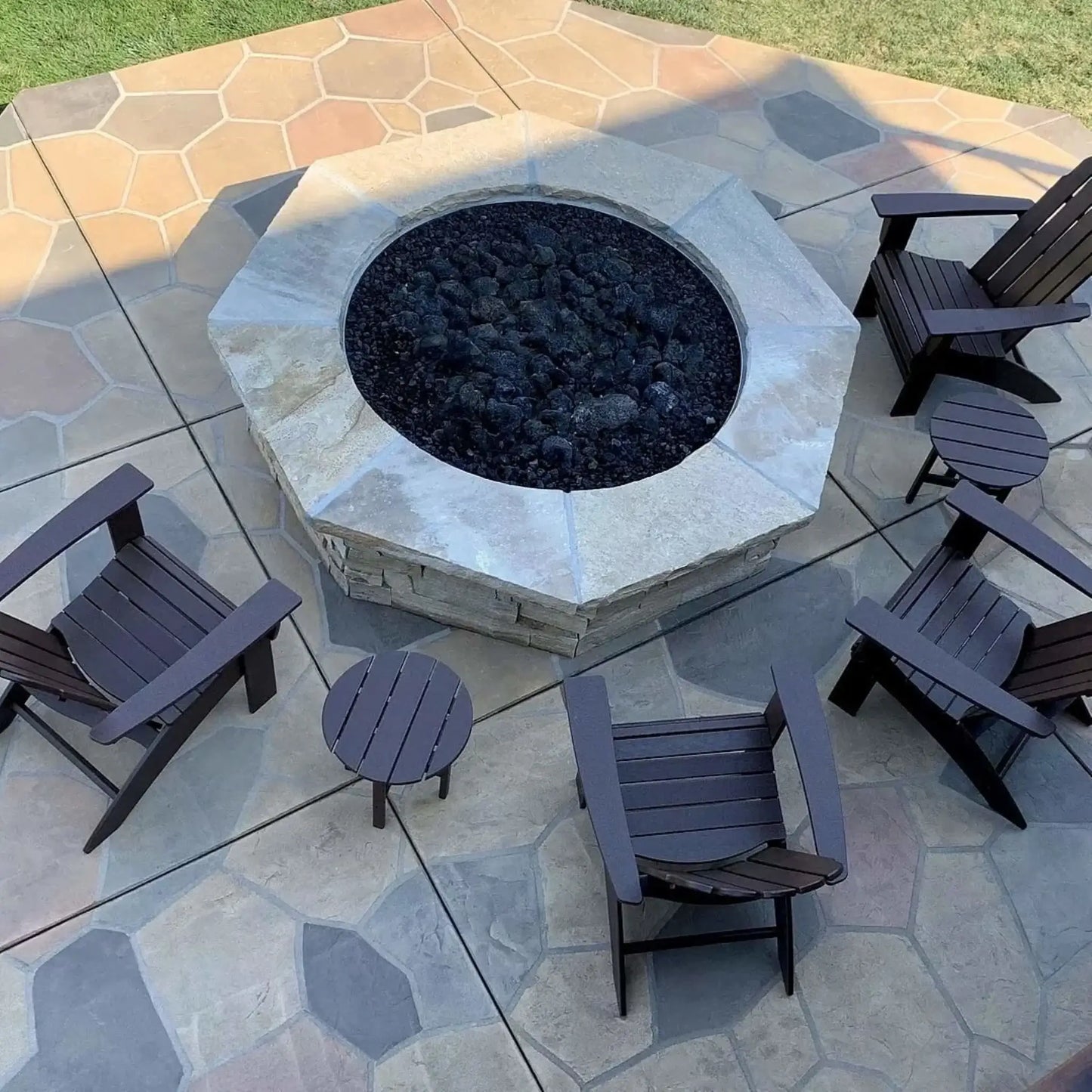 Modern circular concrete fire pit burning with natural gas flames over black lava rock, set on a contemporary patio with wood lounge chairs, side tables, and a warm wood-clad exterior wall for a cozy outdoor living space.