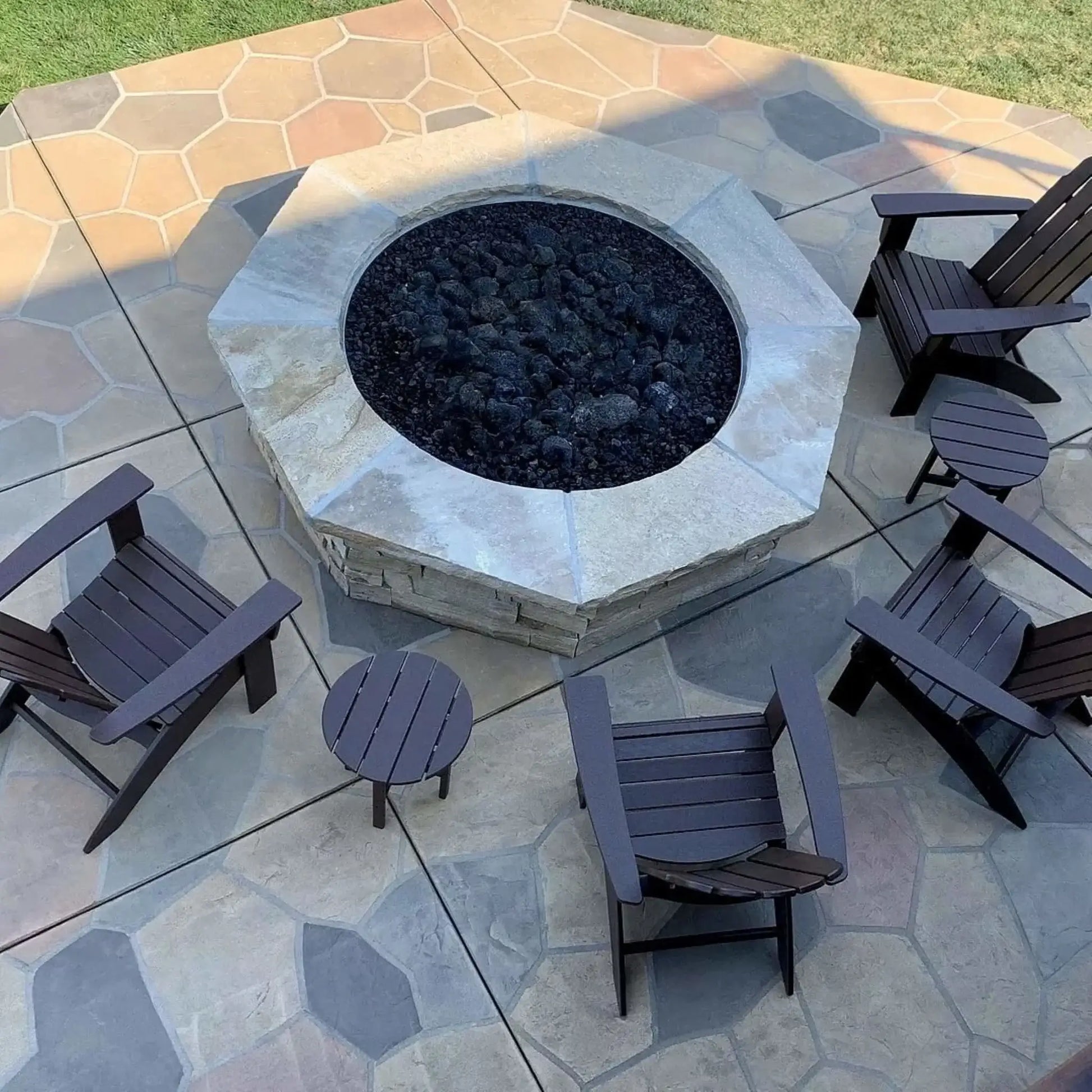 Modern circular concrete fire pit burning with natural gas flames over black lava rock, set on a contemporary patio with wood lounge chairs, side tables, and a warm wood-clad exterior wall for a cozy outdoor living space.
