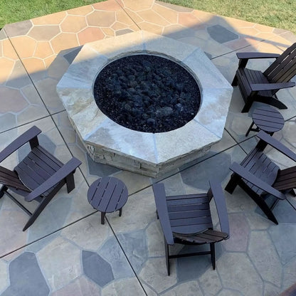Modern circular concrete fire pit burning with natural gas flames over black lava rock, set on a contemporary patio with wood lounge chairs, side tables, and a warm wood-clad exterior wall for a cozy outdoor living space.