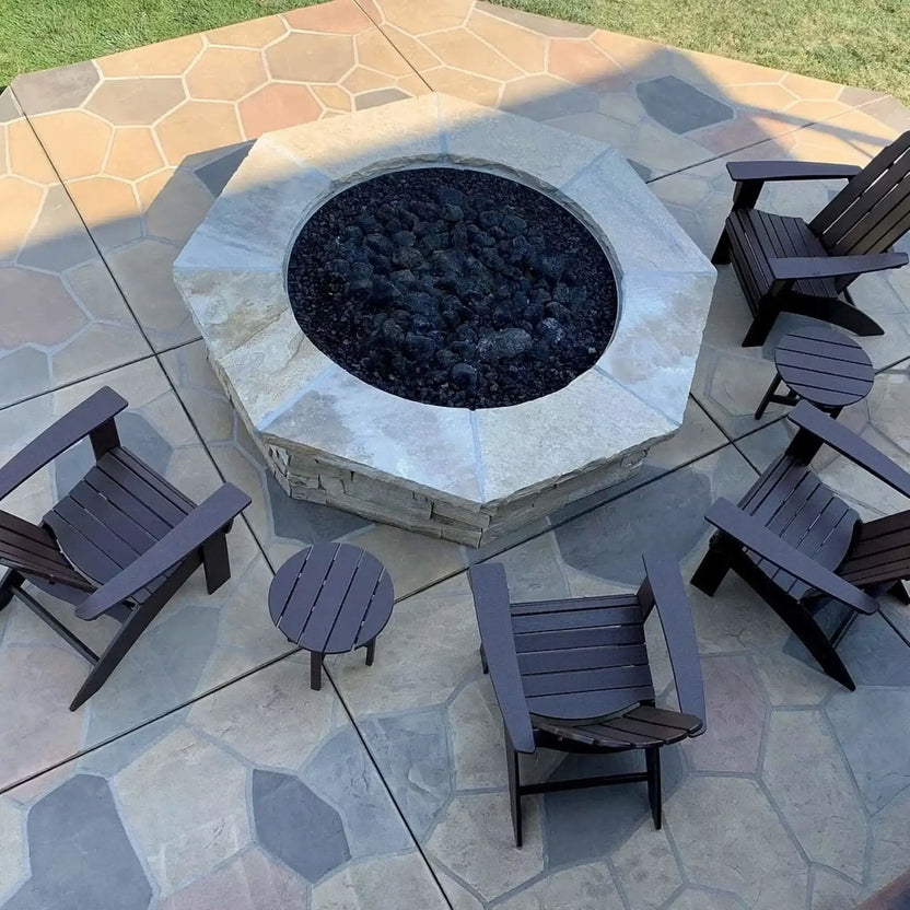 Modern circular concrete fire pit burning with natural gas flames over black lava rock, set on a contemporary patio with wood lounge chairs, side tables, and a warm wood-clad exterior wall for a cozy outdoor living space.