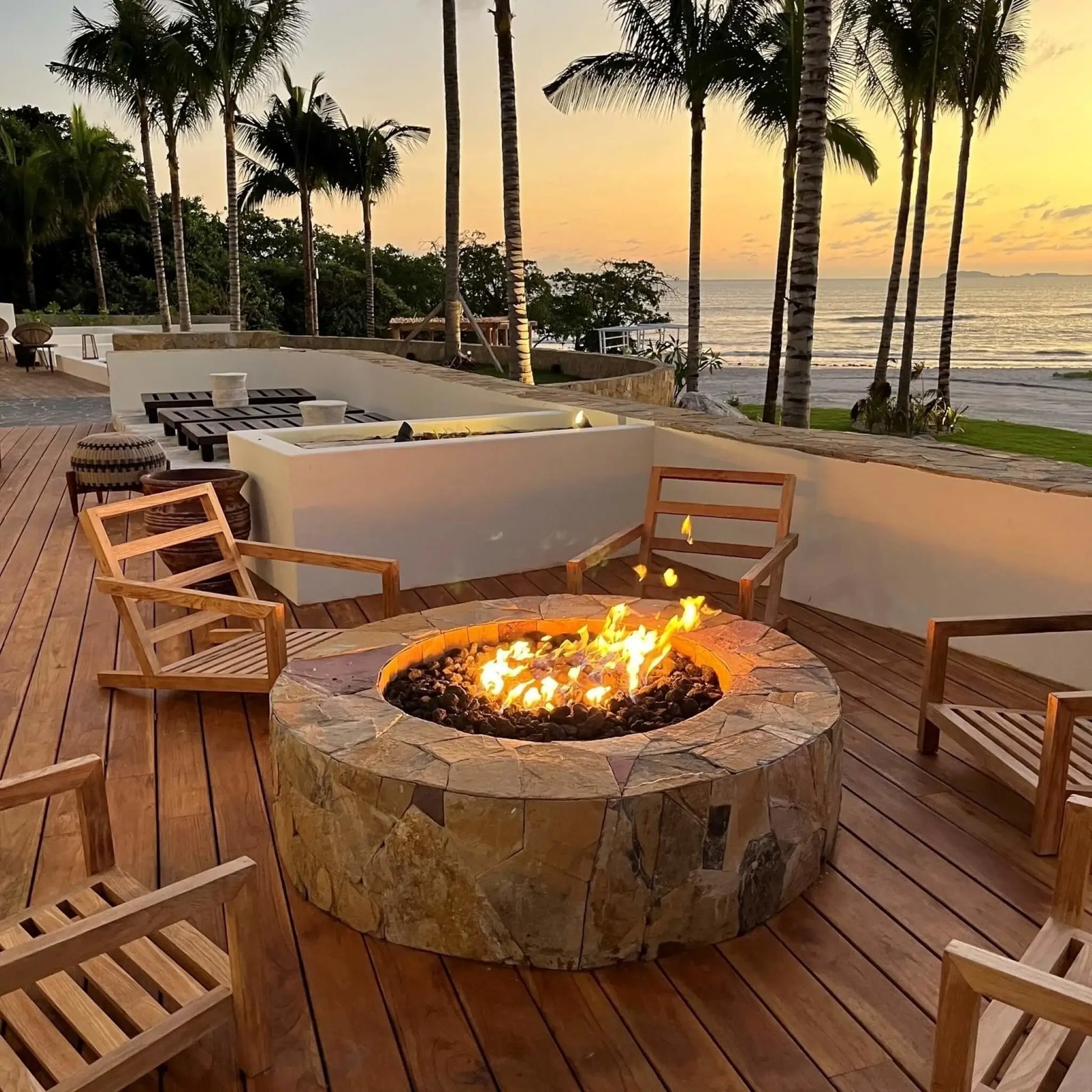Round masonry fire pit filled with black lava rock, centered on a large stone paver patio near a swimming pool, with landscaped greenery, ornamental grasses, and a residential backyard setting in bright daylight.
