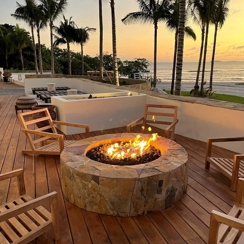 Round masonry fire pit filled with black lava rock, centered on a large stone paver patio near a swimming pool, with landscaped greenery, ornamental grasses, and a residential backyard setting in bright daylight.
