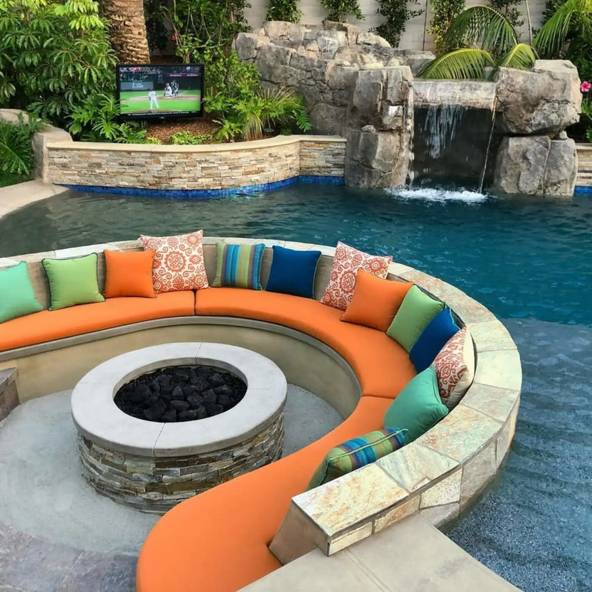 Circular outdoor fire pit with black lava rock, integrated into a curved seating area with colorful cushions beside a resort-style pool and rock waterfall feature, creating a luxury backyard entertaining space.