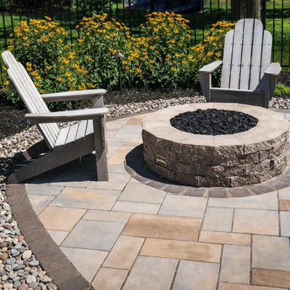 Round stone fire pit with a stainless steel burner pan filled with rolled lava rock, set in a landscaped backyard patio with Adirondack chairs, decorative pavers, and lush greenery creating a cozy outdoor gathering space.