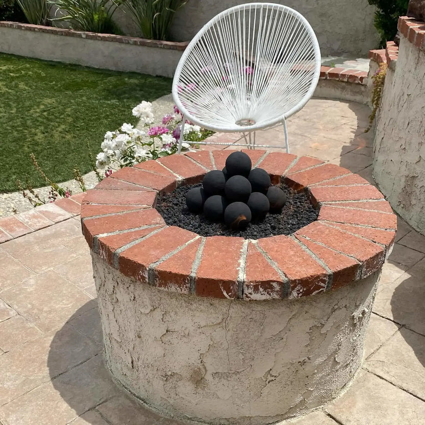 Custom circular fire pit featuring a stainless steel flat burner pan topped with light stone fire balls, surrounded by built-in curved seating and colorful outdoor cushions beside a resort-style pool and waterfall.