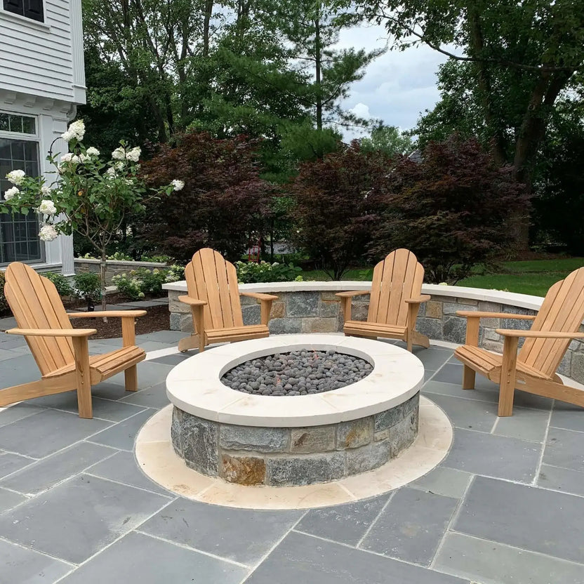 Modern round fire pit constructed with natural stone and a stainless steel burner pan filled with rolled lava stones, positioned on a paved patio overlooking a swimming pool with lounge chairs and landscaped trees.