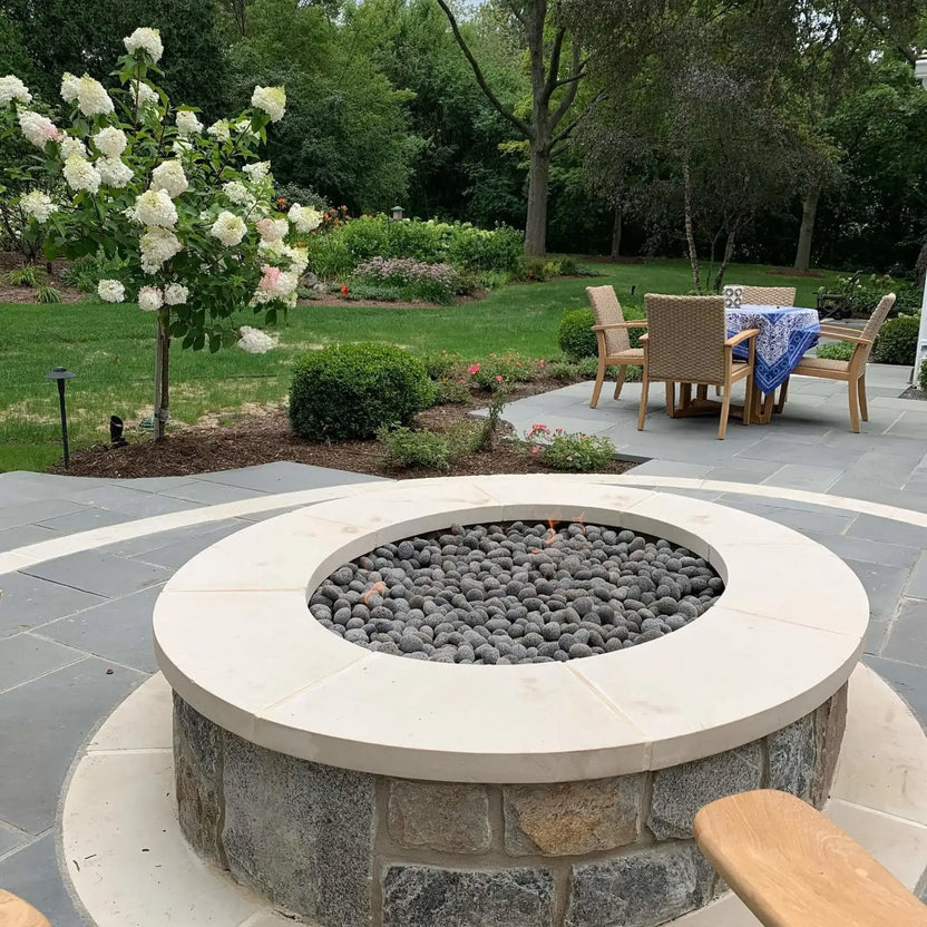 Outdoor circular fire pit with a stainless steel flat burner pan and rolled lava stone media, centered in a contemporary backyard patio with clean paver lines, surrounding seating, and manicured landscaping.