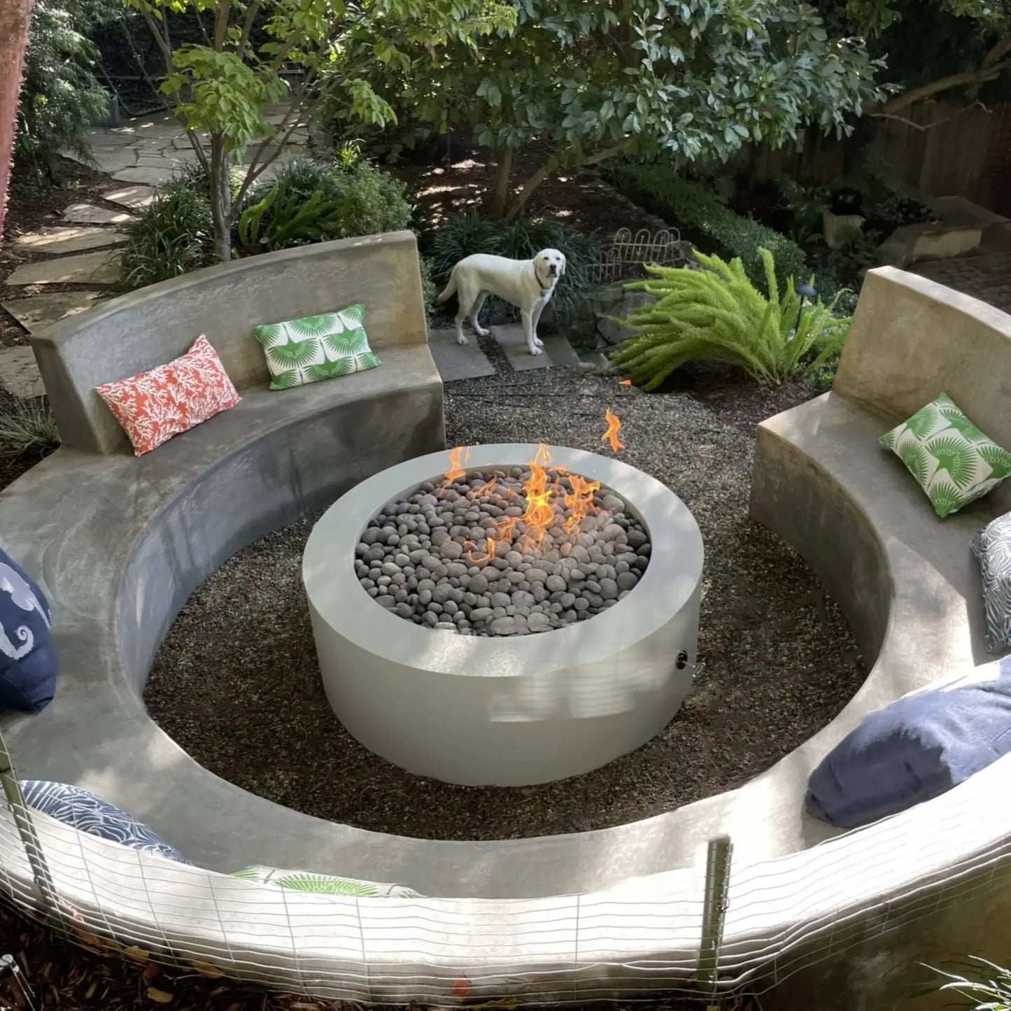 Circular outdoor fire pit with a smooth concrete surround and stacked stone base, filled with rounded lava stones and active flames, centered within curved built-in seating in a landscaped garden setting.