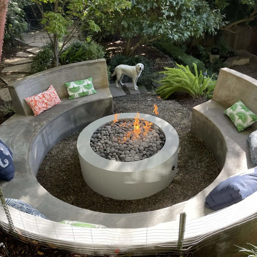Circular outdoor fire pit with a smooth concrete surround and stacked stone base, filled with rounded lava stones and active flames, centered within curved built-in seating in a landscaped garden setting.