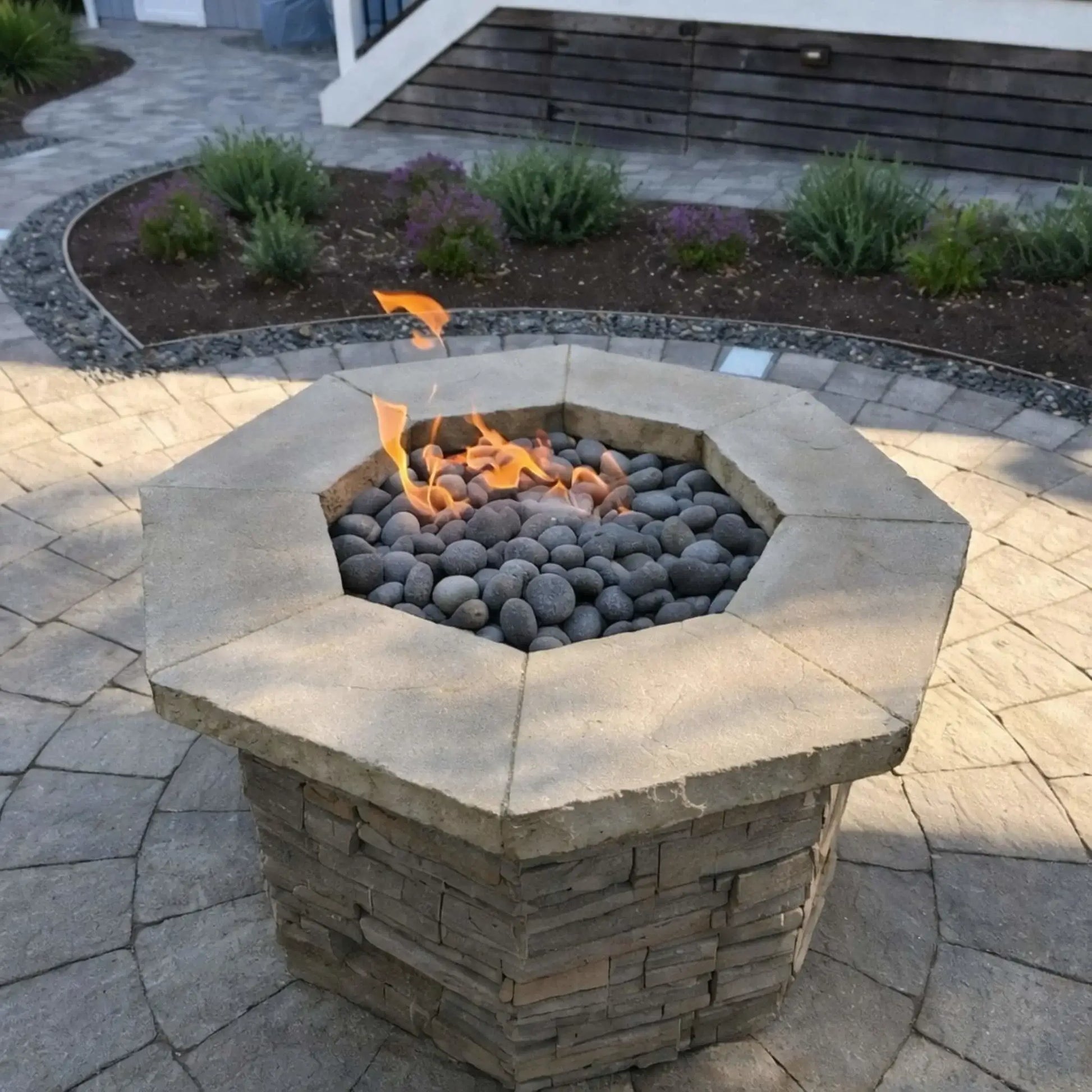 Octagonal stone fire pit with a flat stainless steel burner pan, gray lava stones, and soft flames, installed on a paver patio surrounded by modern landscaping and low garden beds.