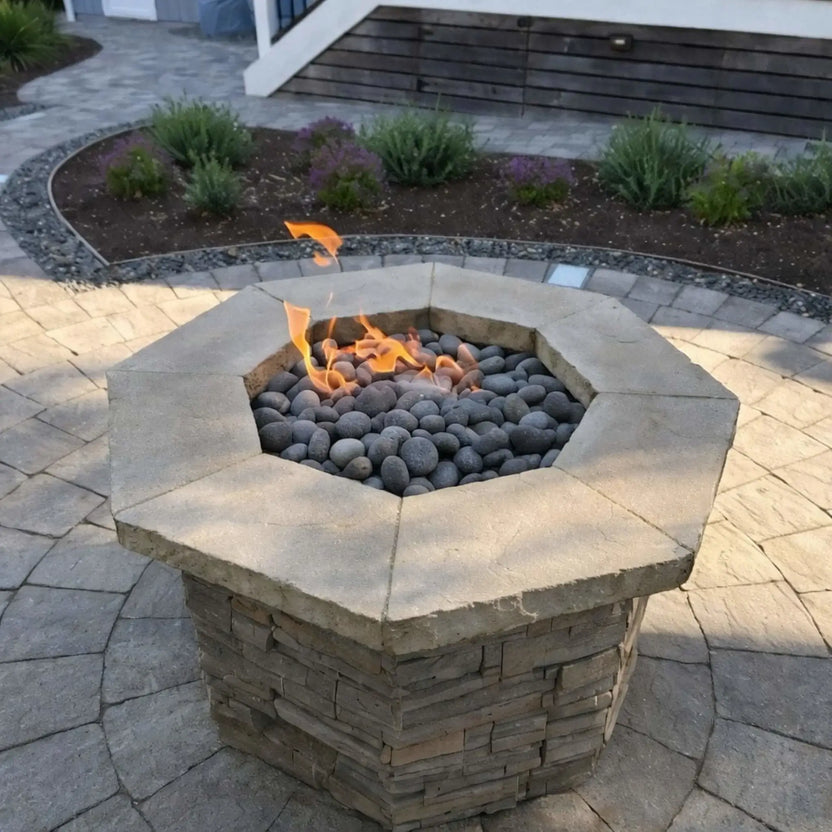 Octagonal stone fire pit with a flat stainless steel burner pan, gray lava stones, and soft flames, installed on a paver patio surrounded by modern landscaping and low garden beds.