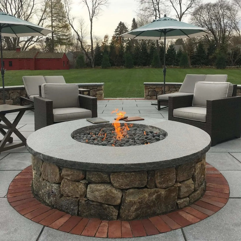 Large round stone fire pit with a wide granite-style coping and stainless steel burner pan, burning over smooth lava stones, positioned between outdoor lounge chairs on a spacious patio with umbrellas and lawn views.