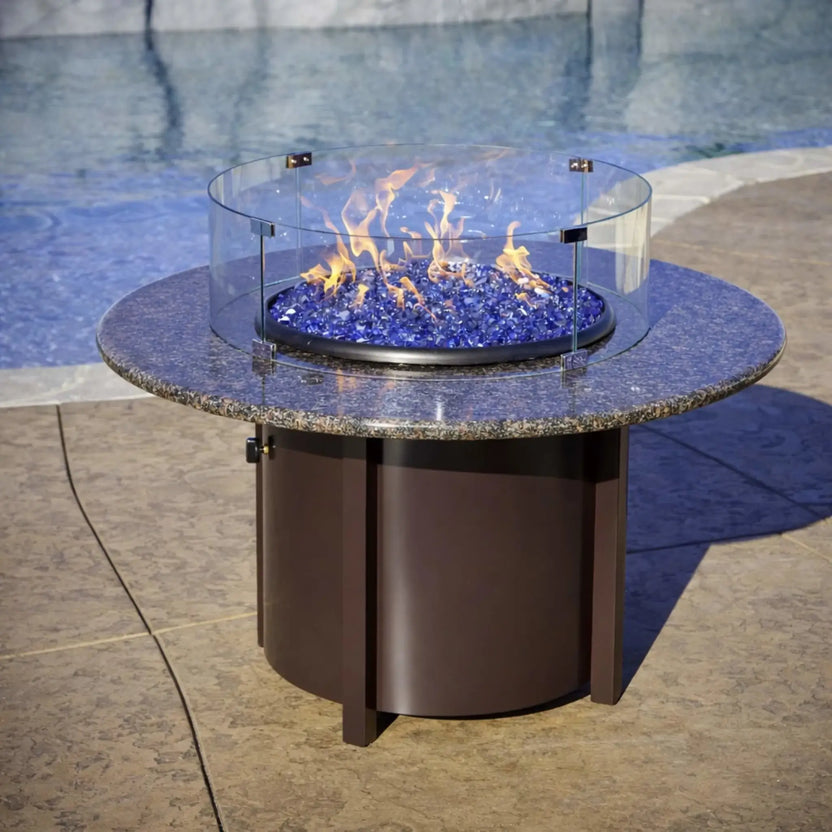 Contemporary round fire pit table with granite top and blue fire glass, featuring a circular glass wind guard that protects the flame while maintaining an open view near a pool deck.
