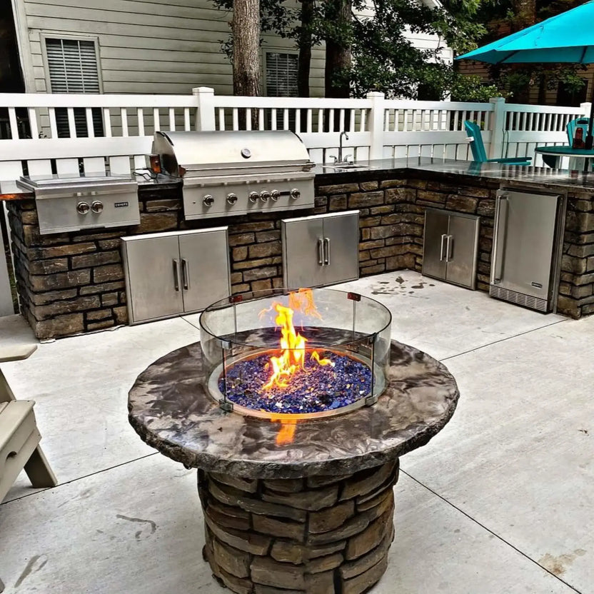 Outdoor round fire pit with textured base and blue fire glass, surrounded by a clear glass wind guard that blocks wind while showcasing tall, steady flames.