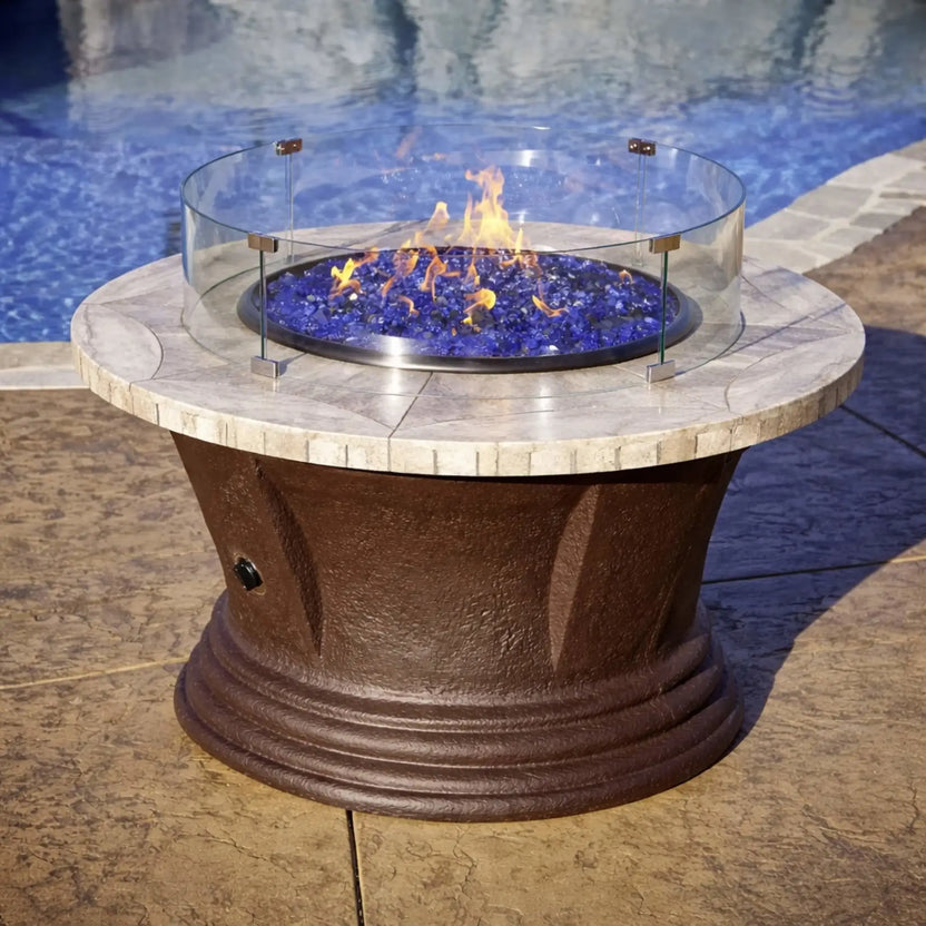 Stone-top round fire pit with vibrant flames and blue fire glass, protected by a circular tempered glass wind guard in a residential backyard patio setting.