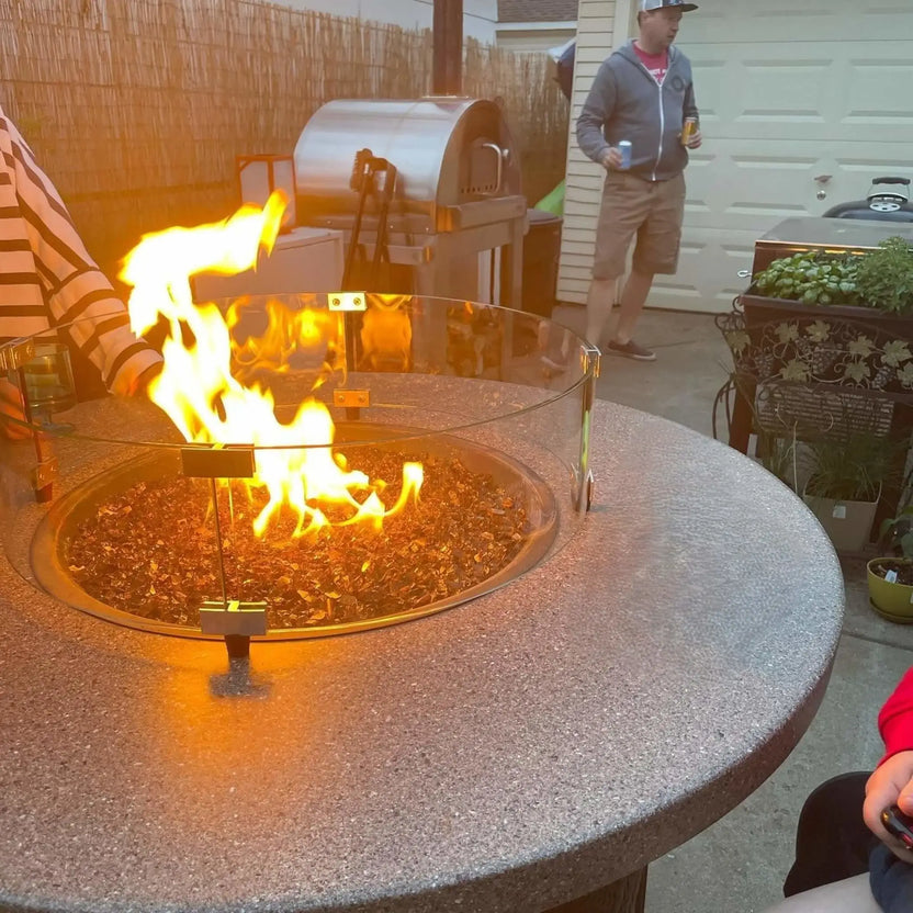 Backyard entertainment area featuring a round fire pit with a glass wind guard, blue fire glass, and active flames, positioned in front of an outdoor kitchen and grill island.