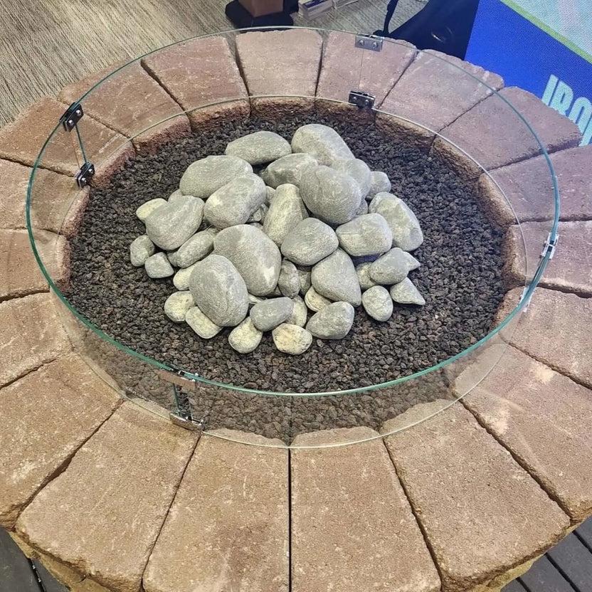 Close-up of a round fire pit filled with natural stone media and lava rock, surrounded by a clear tempered glass wind guard mounted on a stone fire pit enclosure.