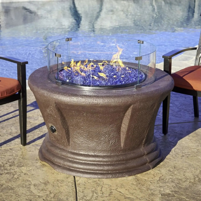 Round fire pit with blue fire glass and active flames, enclosed by a clear tempered glass wind guard, positioned beside a swimming pool for a modern outdoor living setting.