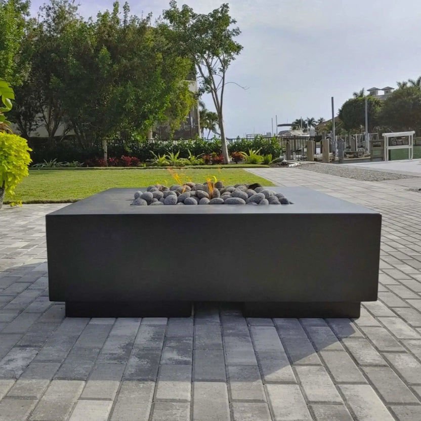Modern square outdoor fire table set on a paved patio near a waterfront property, filled with small lava stones surrounding a low gas flame, with lush landscaping and dock in the background.
